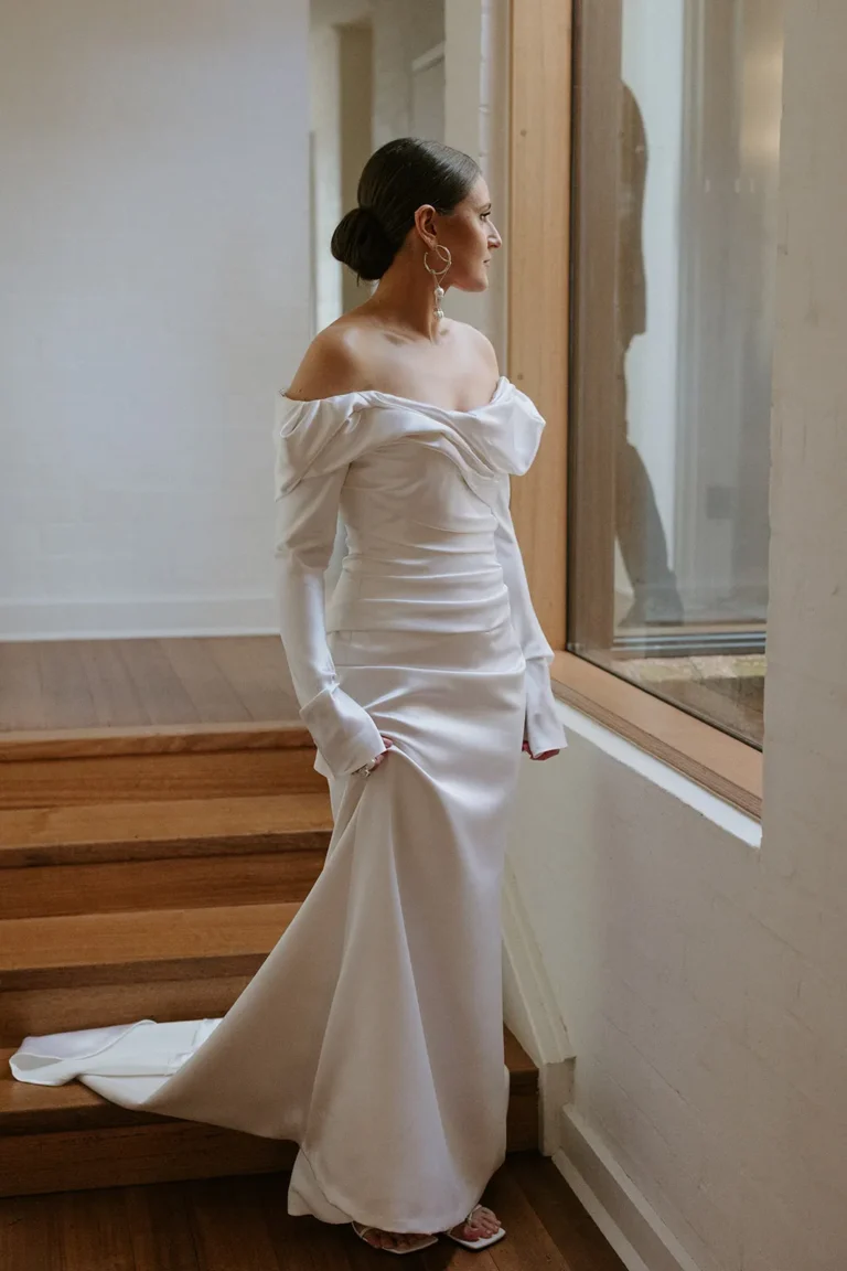 Bride on stairs in satin gown, looking trough the window.