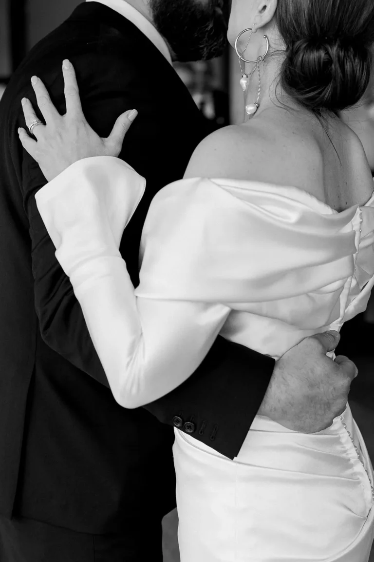 Black-and-white photo of a bride hugging her partner in whide wedding dress and updo hairstyle.