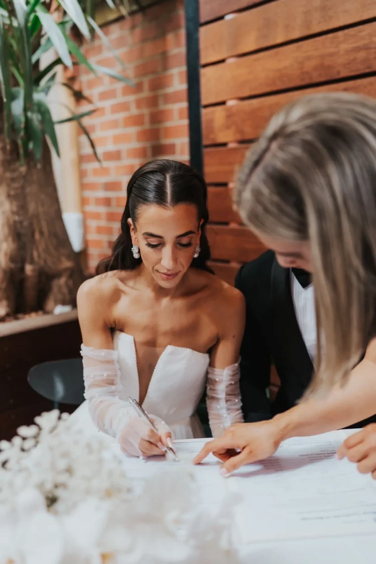 Bride in a strapless wedding dress with sheer, pearl-embellished sleeves signing a document, guided by a woman pointing at the paper, during a wedding ceremony indoors with a wood and brick backdrop.